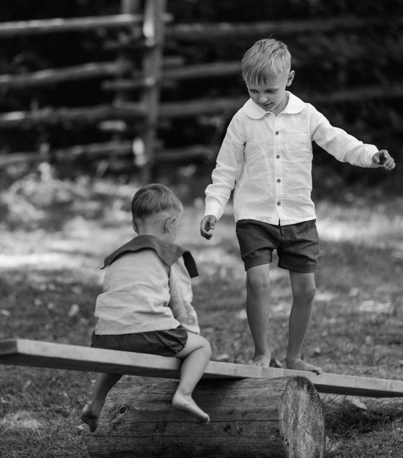 May include: Two young children playing on a seesaw made from a wooden plank and a log. One child is standing, wearing a white button-down shirt and dark shorts. The other child is seated, wearing a sailor-style top and shorts. The scene is in black and white.