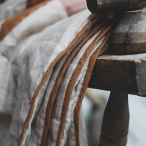 May include: Close-up of a folded linen textile with brown trim, draped over a wooden chair. The fabric is primarily off-white with subtle stripes and a warm, earthy color palette. The chair is made of dark wood.