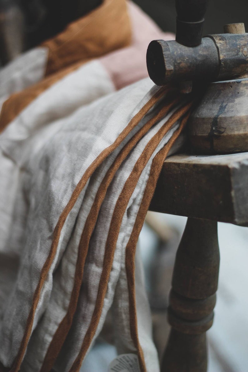 May include: Close-up of a folded linen textile with brown trim, draped over a wooden chair. The fabric is primarily off-white with subtle stripes and a warm, earthy color palette. The chair is made of dark wood.