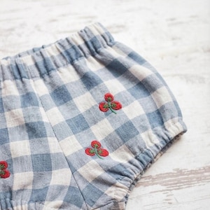 May include: Bloomers featuring a light blue and white gingham check pattern with small embroidered red strawberry motifs. The garment has an elasticated waistband and leg openings, creating a gathered, rounded silhouette throughout the piece.