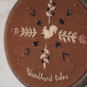 May include: Embroidered hoop art featuring a squirrel, floral, and leaf designs in cream and navy blue thread on a brown fabric. The words "Woodland tales" are stitched in cursive. The wooden hoop is dark brown.