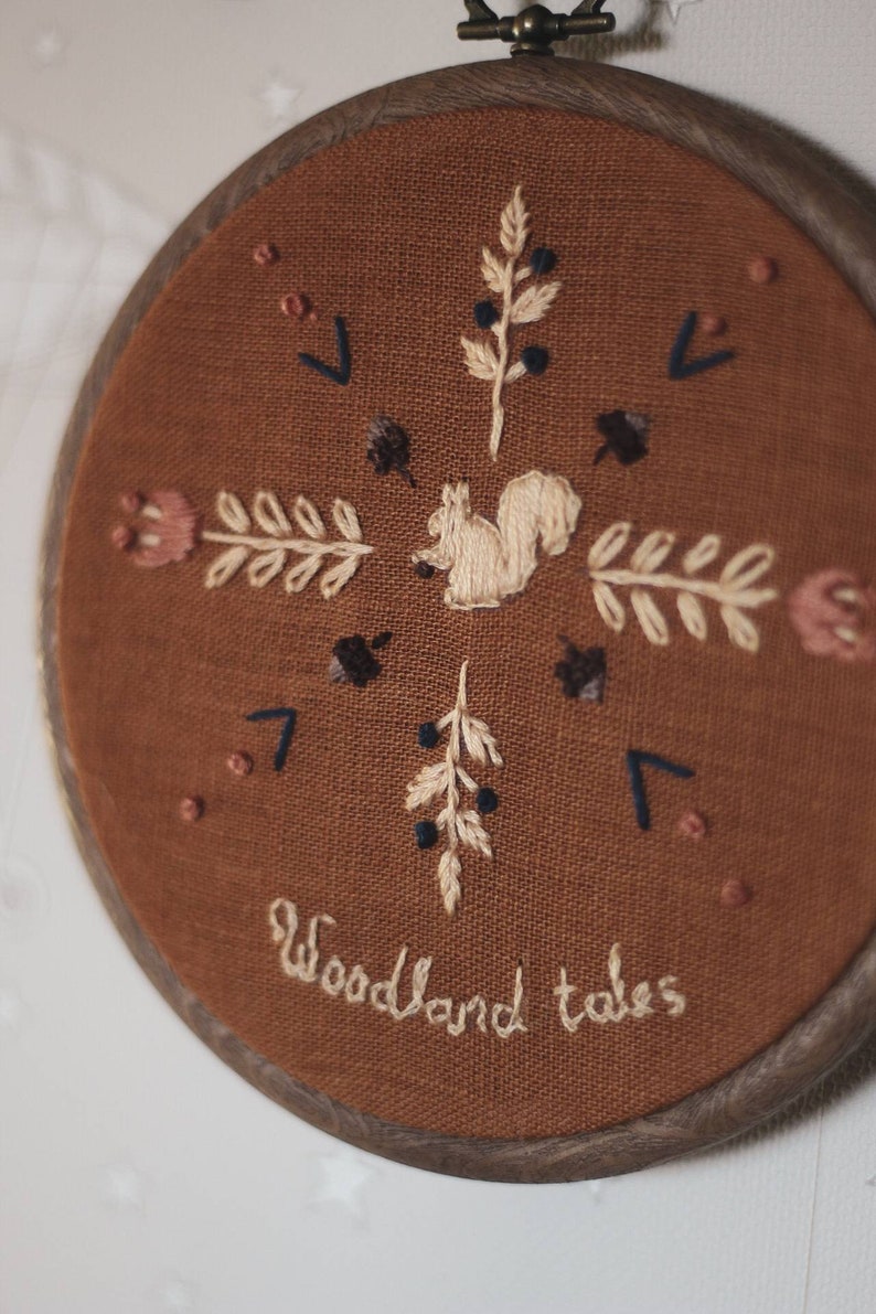 May include: Embroidered hoop art featuring a squirrel, floral, and leaf designs in cream and navy blue thread on a brown fabric. The words "Woodland tales" are stitched in cursive. The wooden hoop is dark brown.