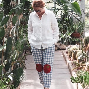 May include: A person wearing a white button-down shirt and blue and white checkered pants with red tomato patches on the knees. The person is standing in a greenhouse with various plants and cacti.