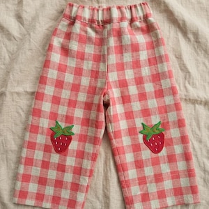 May include: Pants featuring a red and beige gingham check pattern with an elasticized waistband. Each pant leg is decorated with a single embroidered strawberry patch, showcasing a red fruit body with green leaves and small white seed details.