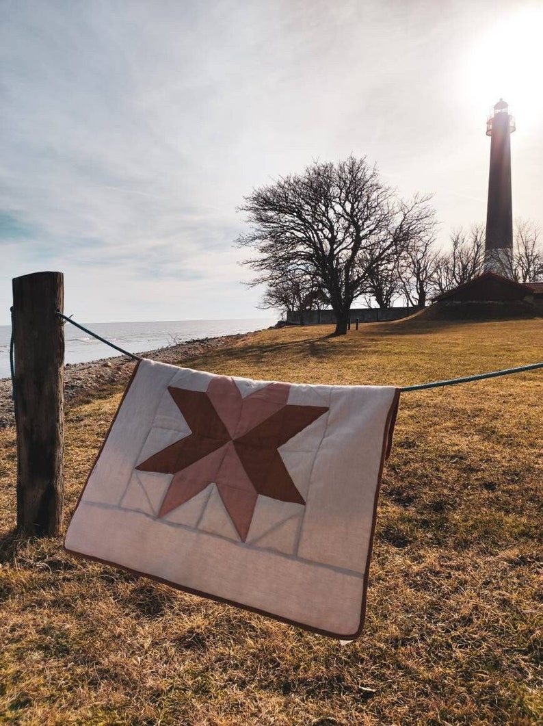 May include: A handmade quilt with a star pattern in shades of brown and pink hangs on a clothesline. The quilt is displayed outdoors with a lighthouse and the ocean in the background. The quilt is a cozy home decor item.