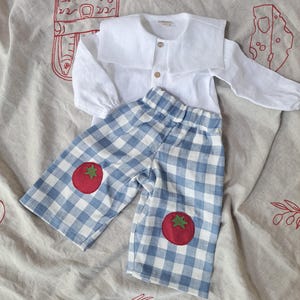Può includere: Una camicia bianca a maniche lunghe con colletto ampio e due bottoni in legno, abbinata a pantaloni a quadri blu e bianchi con due applicazioni a forma di pomodoro rosso con picciolo verde sul davanti, su uno sfondo in tessuto beige.