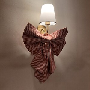 May include: A wall-mounted lamp with a white lampshade and a gold-colored base. The lamp is adorned with a large, decorative bow made of red and white striped fabric, adding a whimsical touch to the design.
