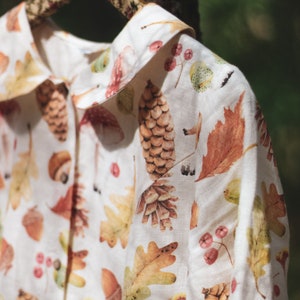 Puede incluir: Una camisa de lino blanca con un estampado de oto&ntilde;o que incluye setas, bellotas, hojas y pi&ntilde;as.