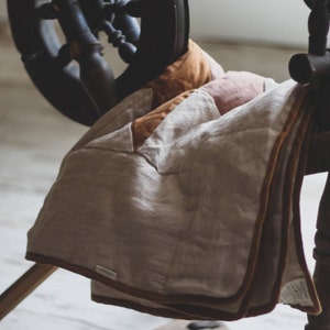 May include: A neutral-toned linen blanket with a brown border is draped over a wooden spinning wheel. The blanket has a patchwork design with shades of beige, tan, and brown. The spinning wheel is dark wood, and the background is a soft, neutral color.