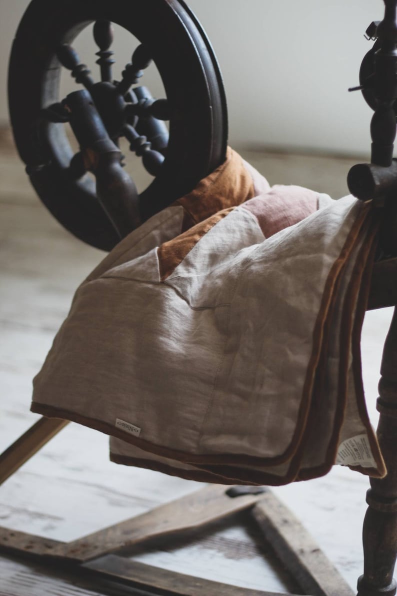 May include: A neutral-toned linen blanket with a brown border is draped over a wooden spinning wheel. The blanket has a patchwork design with shades of beige, tan, and brown. The spinning wheel is dark wood, and the background is a soft, neutral color.