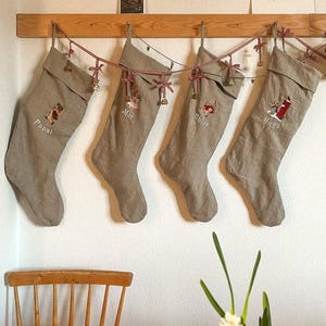 Può includere: Quattro calze di Natale beige con nastro rosso e bianco e campane appese a uno scaffale di legno. Le calze sono ricamate con i nomi "Papà", "Mamma", "Chloe" e "Hugo".