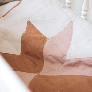 May include: A handmade quilt with a starburst pattern in shades of cream, light pink, and brown. The quilt is made of fabric and is placed in a white crib. The quilt is a soft, cozy, and decorative item.