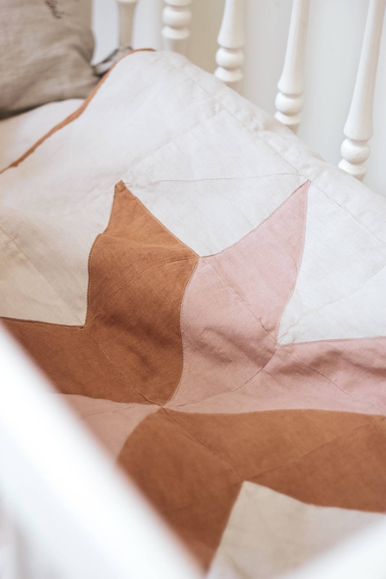 May include: A handmade quilt with a starburst pattern in shades of cream, light pink, and brown. The quilt is made of fabric and is placed in a white crib. The quilt is a soft, cozy, and decorative item.