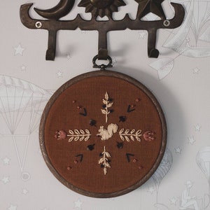 May include: A decorative wall hook with celestial and star motifs, holding a round, embroidered artwork. The artwork features a squirrel and floral design on a brown background, framed in wood.