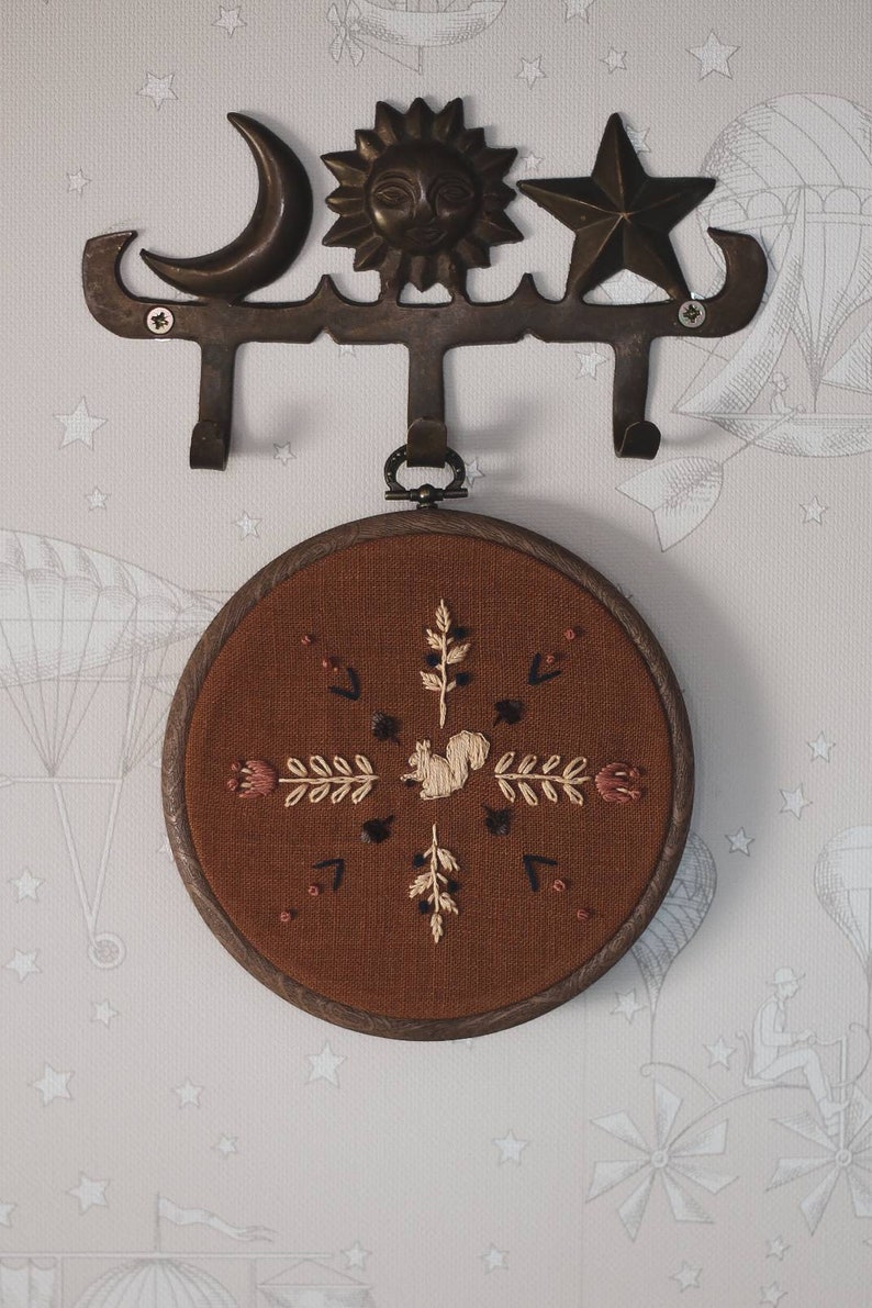 May include: A decorative wall hook with celestial and star motifs, holding a round, embroidered artwork. The artwork features a squirrel and floral design on a brown background, framed in wood.