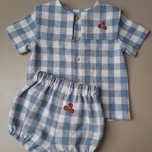 Op de afbeelding: Een blauw en wit geruit baby-outfit, bestaande uit een shirt met korte mouwen en bijpassende bloeiers. Het shirt heeft een ronde kraag en houten knopen. Beide stukken zijn geborduurd met een klein rood bloemenmotief.