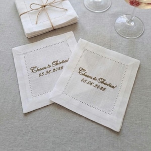 Pode incluir: Guardanapos de cocktail de linho branco com bordado "Cheers to Thirties!" e a data 05.06.2026. Uma pilha de guardanapos amarrados com barbante é visível. Os guardanapos são quadrados com uma borda pontilhada.