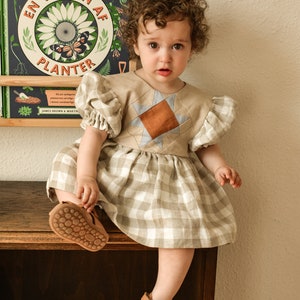 Può includere: Una bambina che indossa un vestito a quadri bianco e marrone chiaro con un design patchwork blu e marrone sul corpetto. La bambina è seduta su uno scaffale di legno con un libro intitolato "En Verden Af Planter" dietro di lei.