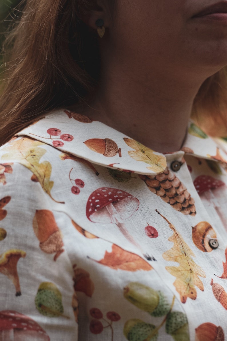 Puede incluir: Una camisa de lino blanca con cuello de Peter Pan con un estampado de bosque de setas, bellotas, hojas y bayas.