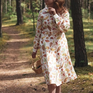 Puede incluir: Una mujer con un vestido blanco con estampado de oto&ntilde;o camina por un sendero en el bosque. El vestido tiene hojas, bellotas y pi&ntilde;as. Lleva una cesta de mimbre.