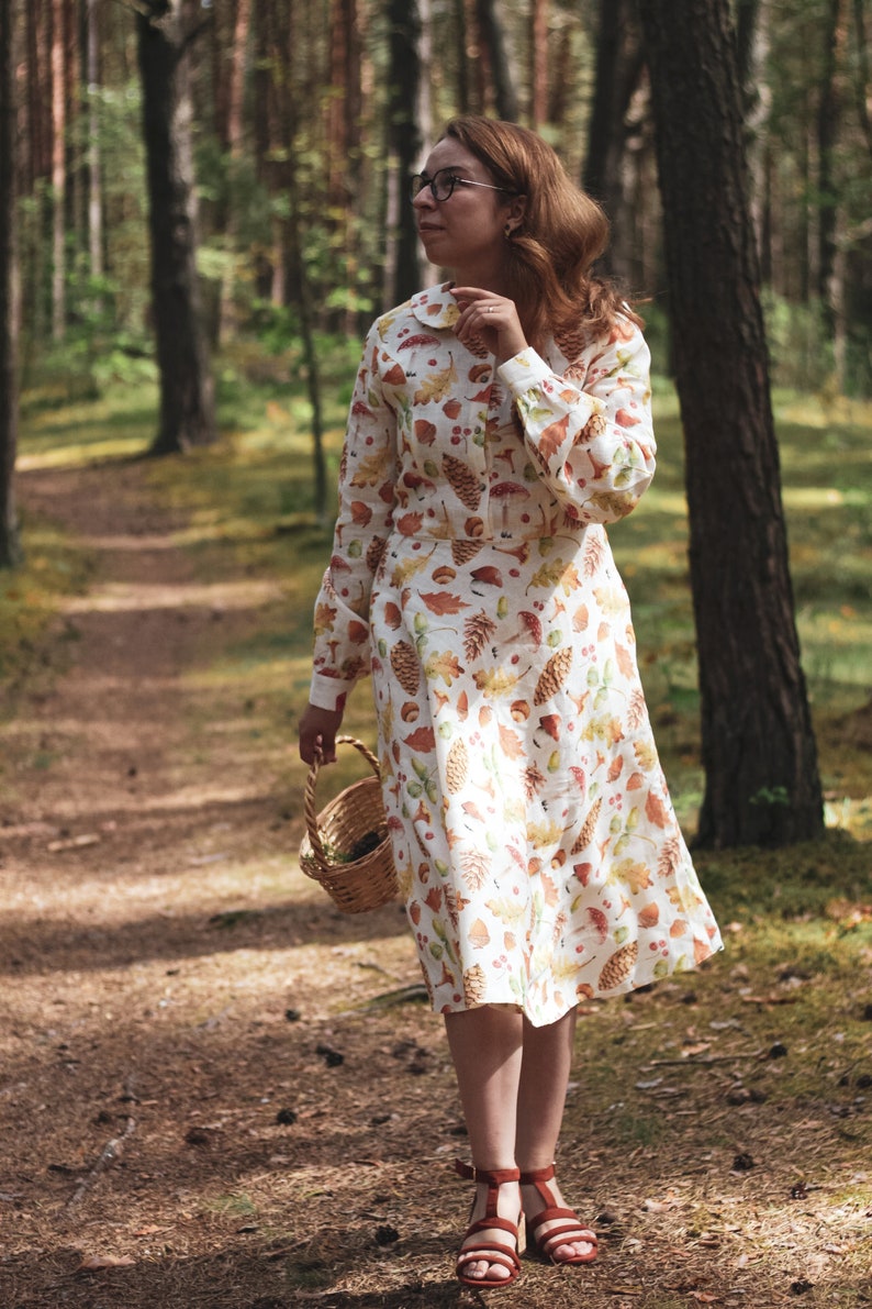 Puede incluir: Una mujer con un vestido blanco con estampado de oto&ntilde;o camina por un sendero en el bosque. El vestido tiene hojas, bellotas y pi&ntilde;as. Lleva una cesta de mimbre.