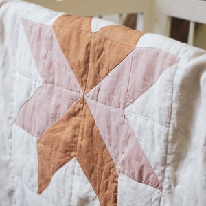 May include: A handmade quilt featuring a star design in brown and pink hues. The star is set against a cream backdrop. The quilt is draped over a white cot.