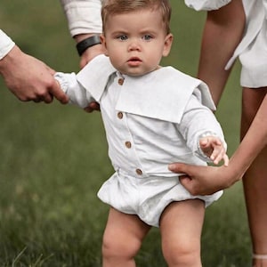 May include: A baby wearing a white linen romper with a Peter Pan collar and wooden buttons. The baby is standing on green grass, with hands holding the baby's arms and waist.
