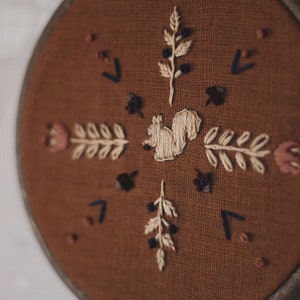May include: Embroidered wall art featuring a squirrel and botanical elements. The design is stitched on a brown fabric, set within a round wooden hoop. The embroidery includes beige, brown, and dark blue details, creating a rustic, nature-inspired aesthetic.