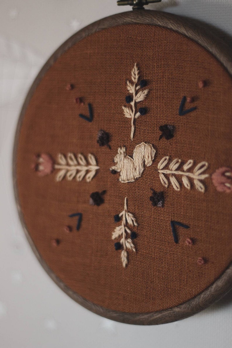 May include: Embroidered wall art featuring a squirrel and botanical elements. The design is stitched on a brown fabric, set within a round wooden hoop. The embroidery includes beige, brown, and dark blue details, creating a rustic, nature-inspired aesthetic.