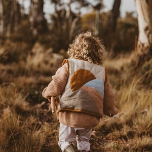 Puede incluir: Un niño lleva un chaleco acolchado marrón y blanco con un diseño de arcoíris y sol caminando por un campo de hierba alta. El chaleco tiene un ribete marrón y un diseño de patchwork azul claro, rosa y naranja.