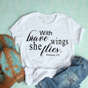 Könnte beinhalten: Weißes T-Shirt mit dem Aufdruck "With brave wings she flies. Philippians 4:13". Das Shirt liegt auf einem hellblauen Untergrund mit einem Paar braunen Cowboystiefeln und einer blauen Jeans.
