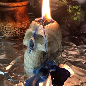 May include: A white skull-shaped candle with blue and black wax drippings, burning with a bright orange flame. The candle is sitting on a silver foil surface. The candle has the words "HOODOO" and "ROOT" written on it.