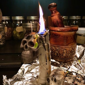 Puede incluir: Una vela blanca con forma de calavera con una llama azul que arde en la parte superior. La vela está sobre una superficie de papel de aluminio plateado junto a un mortero y un pilón de madera y varios frascos de vidrio.