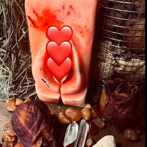 Puede incluir: Una vela rosa y roja con un diseño de tres corazones. La vela está rodeada de pétalos de rosa, cristales y piedras de cornalina.