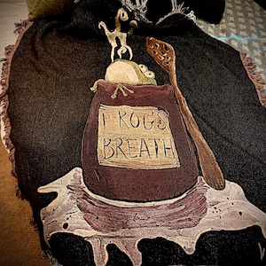 Può includere: Coperta nera tessuta con un design stravagante. La coperta presenta una sacca marrone con le parole "FROGS BREATH" e una rana dei cartoni animati. Sono raffigurati anche un cucchiaio di legno e una piccola creatura. La coperta ha un bordo sfrangiato.