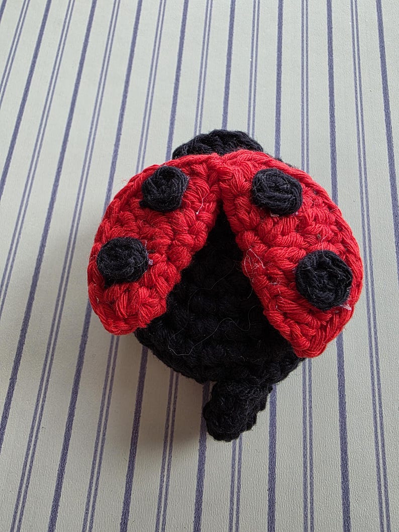 May include: A handmade crochet ladybug with a red body and black spots. The ladybug's head and underside are black. The wings are open, showcasing the detailed crochet work. This whimsical item is likely a decorative piece or a toy.