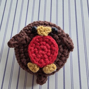 May include: A handmade, crocheted robin bird with a round body. The bird features a brown body, a red breast, yellow accents, and black bead eyes. The bird is sitting on a striped background.