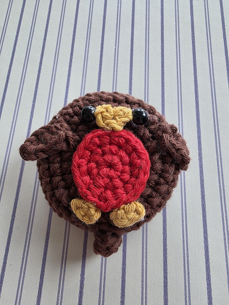 May include: A handmade, crocheted robin bird with a round body. The bird features a brown body, a red breast, yellow accents, and black bead eyes. The bird is sitting on a striped background.
