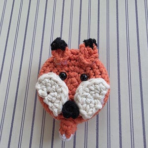 May include: A crocheted fox head with orange, black, and white yarn. The fox has black eyes, black ear tips, and white accents around the nose and mouth. The fox is a handmade item.