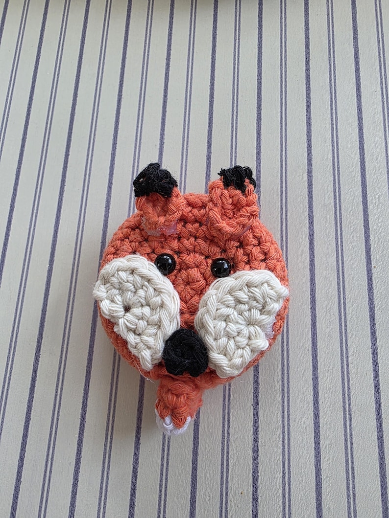 May include: A crocheted fox head with orange, black, and white yarn. The fox has black eyes, black ear tips, and white accents around the nose and mouth. The fox is a handmade item.
