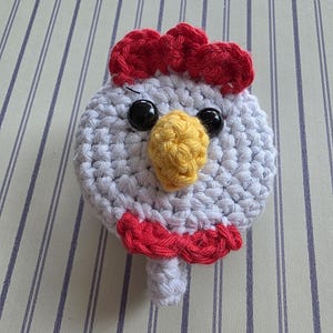 May include: A handmade crochet chicken head with a white body, red comb and wattle, yellow beak, and black eyes. The chicken is made of yarn and has a whimsical, cartoonish appearance. The craft item is likely a decorative piece or toy.