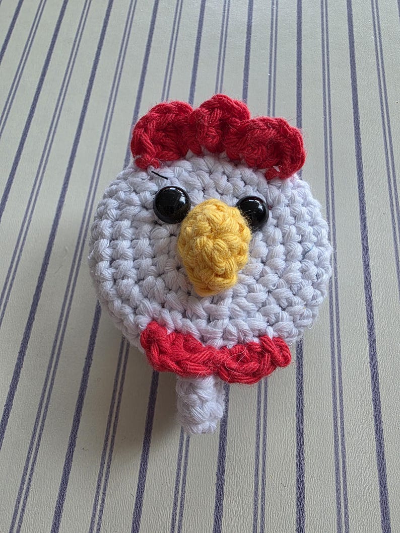 May include: A handmade crochet chicken head with a white body, red comb and wattle, yellow beak, and black eyes. The chicken is made of yarn and has a whimsical, cartoonish appearance. The craft item is likely a decorative piece or toy.