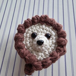 May include: A handmade crochet lion face with a cream-colored center and a brown mane. The lion has black button eyes and a black nose. The item is likely a decorative piece or a toy.
