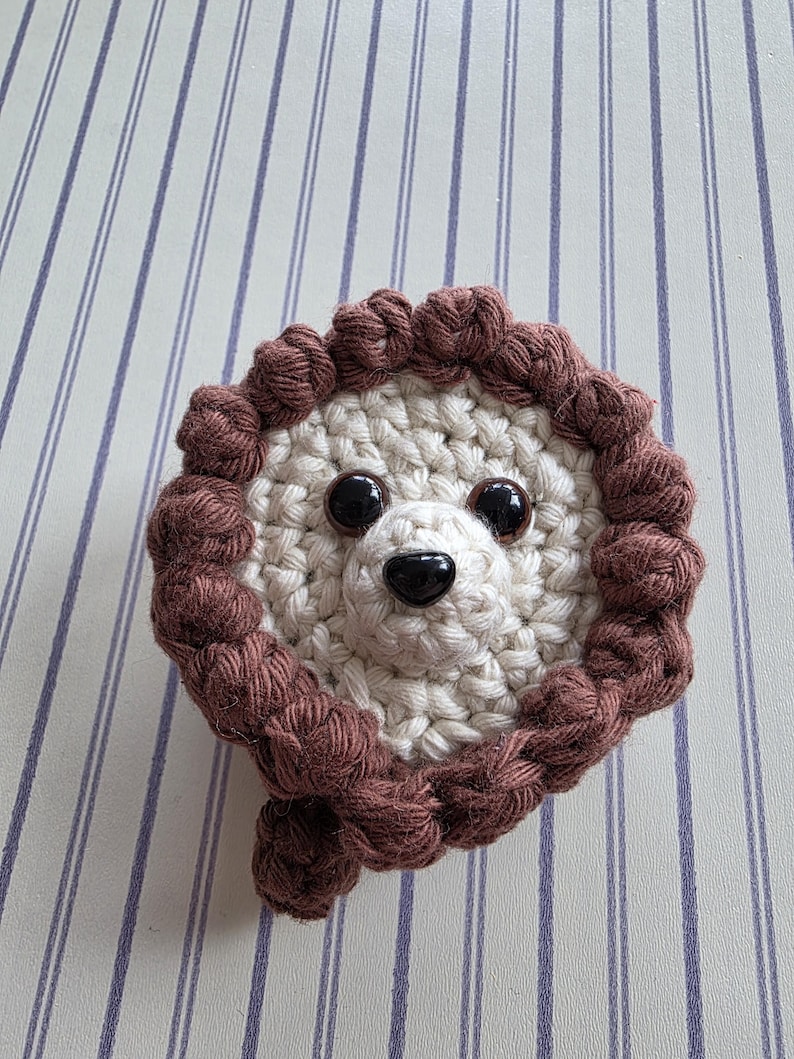 May include: A handmade crochet lion face with a cream-colored center and a brown mane. The lion has black button eyes and a black nose. The item is likely a decorative piece or a toy.