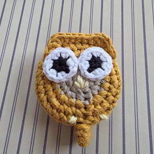 May include: A handmade, crocheted owl in shades of mustard yellow, white, gray, and black. The owl has large, round eyes with white and black details. The owl's body is a rounded shape with a small, pointed beak and a small tail.