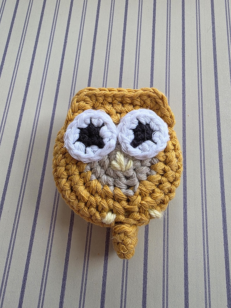 May include: A handmade, crocheted owl in shades of mustard yellow, white, gray, and black. The owl has large, round eyes with white and black details. The owl's body is a rounded shape with a small, pointed beak and a small tail.