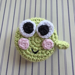 May include: A handmade, crocheted green frog face with large white-rimmed eyes, black pupils, and pink cheeks. The frog has a small, curved mouth and a small, green appendage. The frog is made of yarn.