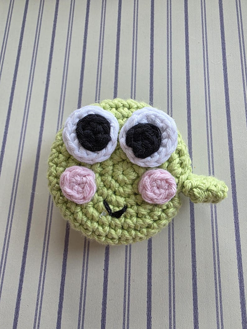 May include: A handmade, crocheted green frog face with large white-rimmed eyes, black pupils, and pink cheeks. The frog has a small, curved mouth and a small, green appendage. The frog is made of yarn.