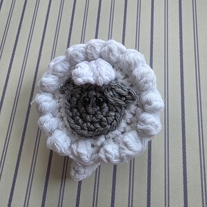 May include: A handmade crochet sheep coaster. The coaster features a white, fluffy wool-like texture around the perimeter, with a gray face and black eyes in the center. The sheep has a small, rounded head and a fluffy body.