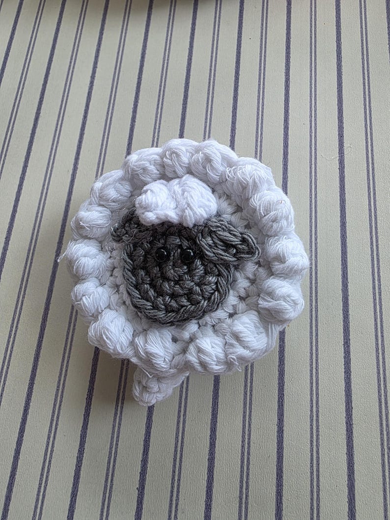 May include: A handmade crochet sheep coaster. The coaster features a white, fluffy wool-like texture around the perimeter, with a gray face and black eyes in the center. The sheep has a small, rounded head and a fluffy body.